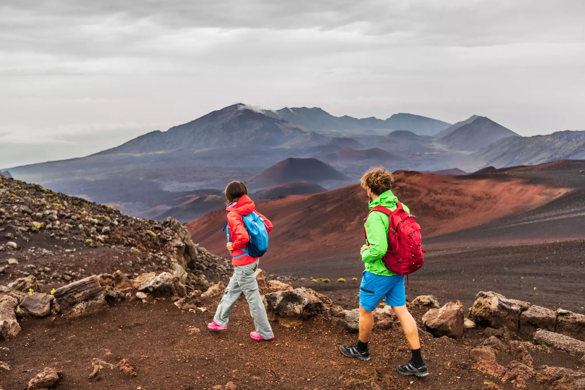 Two people one in a red jacket, the other in a green jacket, carrying backpacks hike on a volcano with Haleakalā volcano in the background.