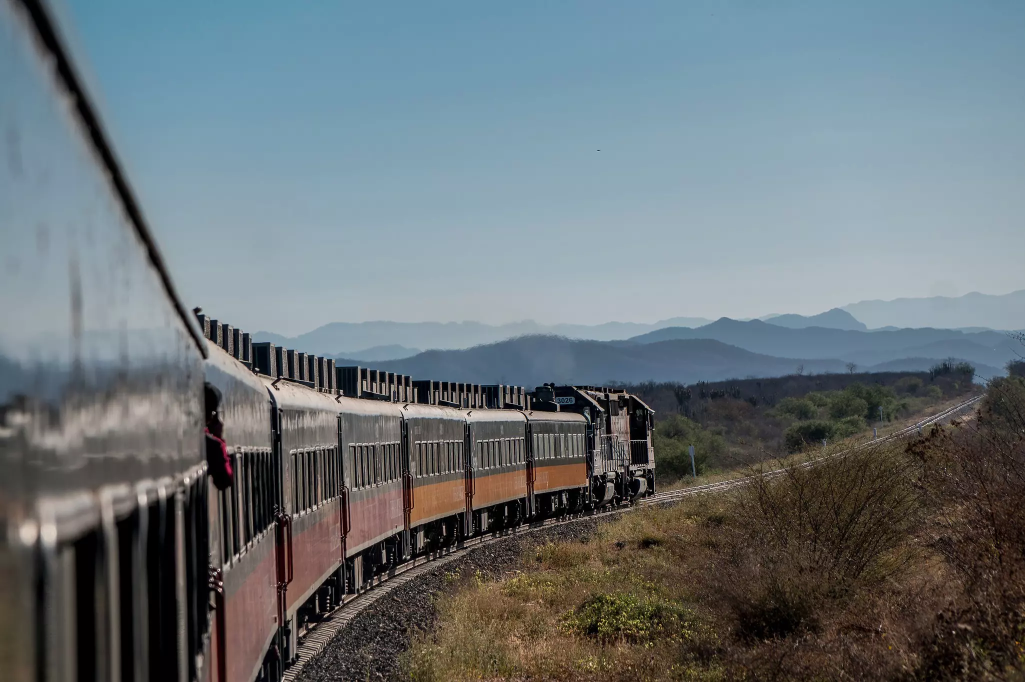 Part of a train in close-up to the left curving around tracks with hazy mountains in the distance on a partly sunn day.