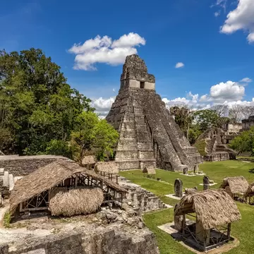 Thatched shelters over ancient stone ruins and upright stelae on a lawn, a steep stepped pyramid rising behind