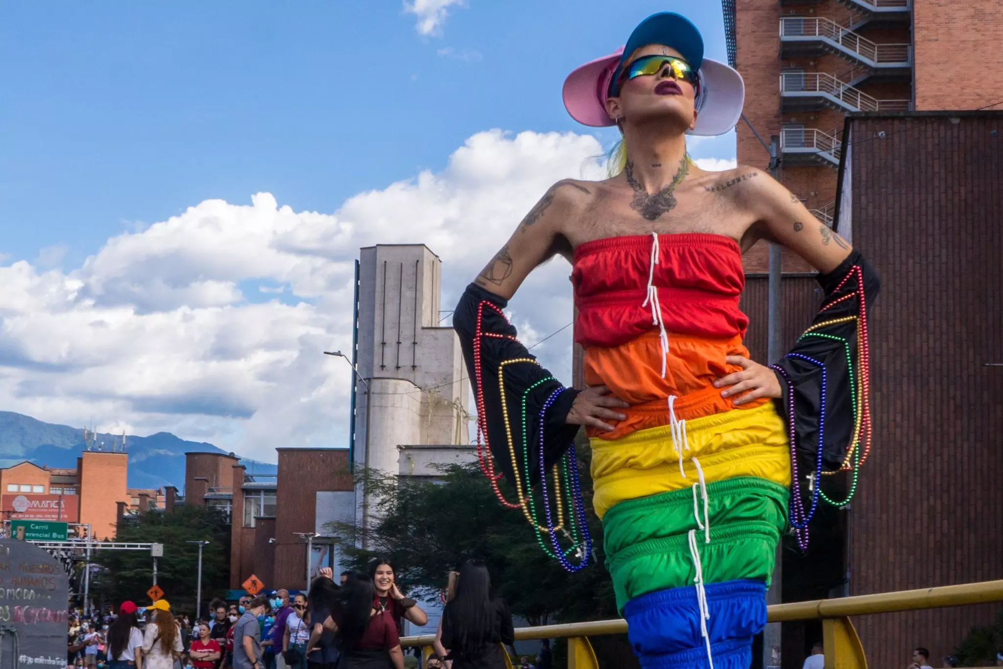 A drag queen dressed in a colorful rainbow-patterned dress draped with beads strikes a pose at an outdoor Pride event in a city