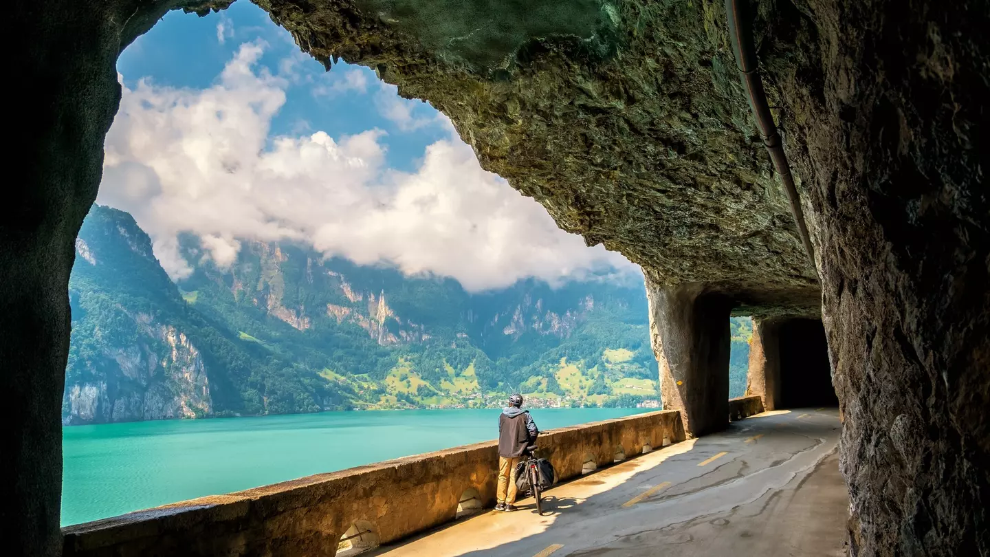 A man stands next to his bike in a tunnel, facing a lake and a mountain range.
