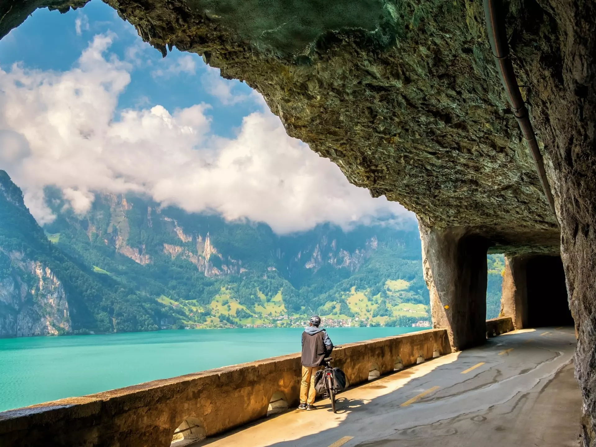 A man stands next to his bike in a tunnel, facing a lake and a mountain range.