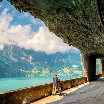 A man stands next to his bike in a tunnel, facing a lake and a mountain range.