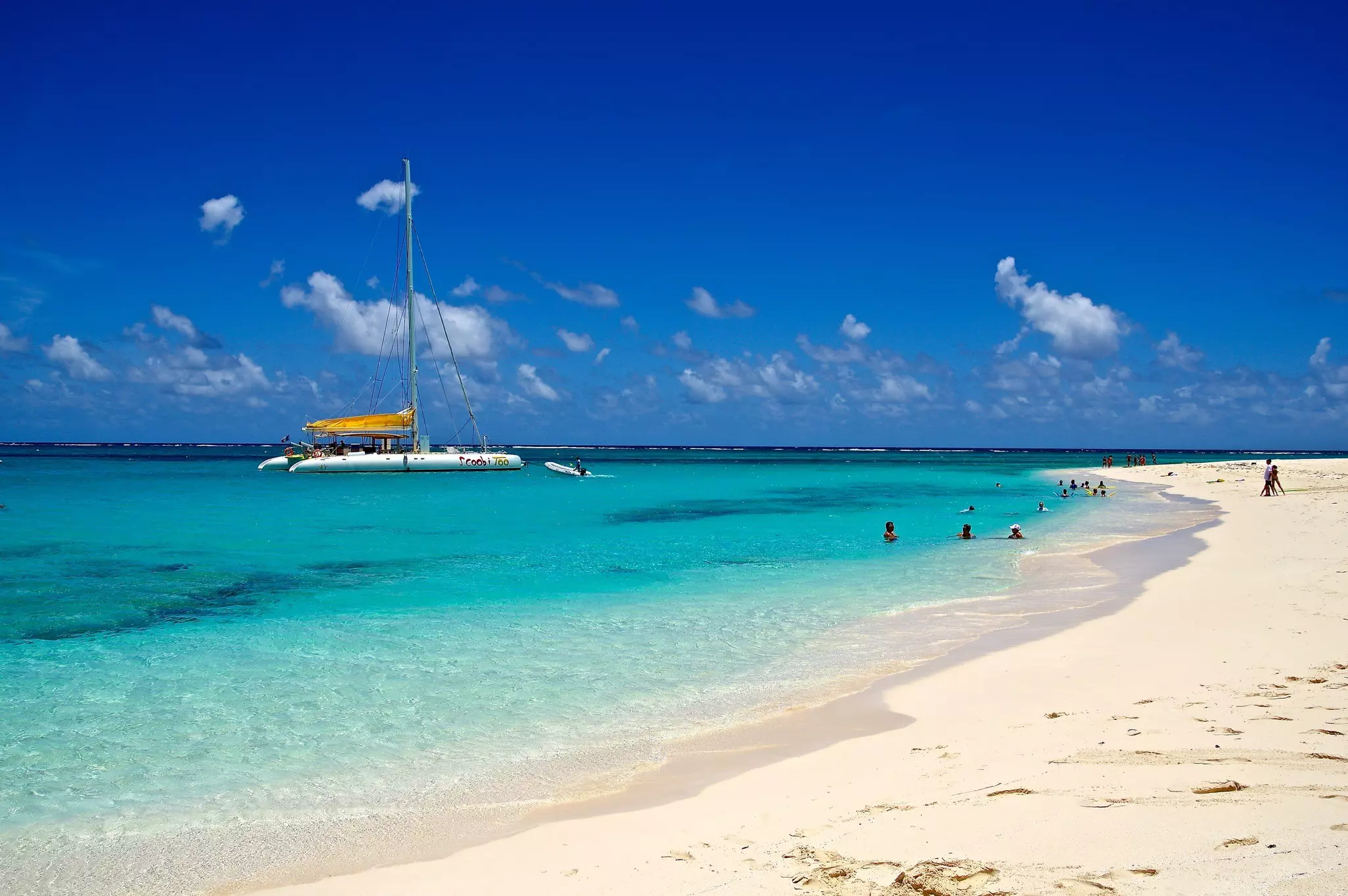 A catamaran is moored in the shallow azure waters off a wide tropical beach.
