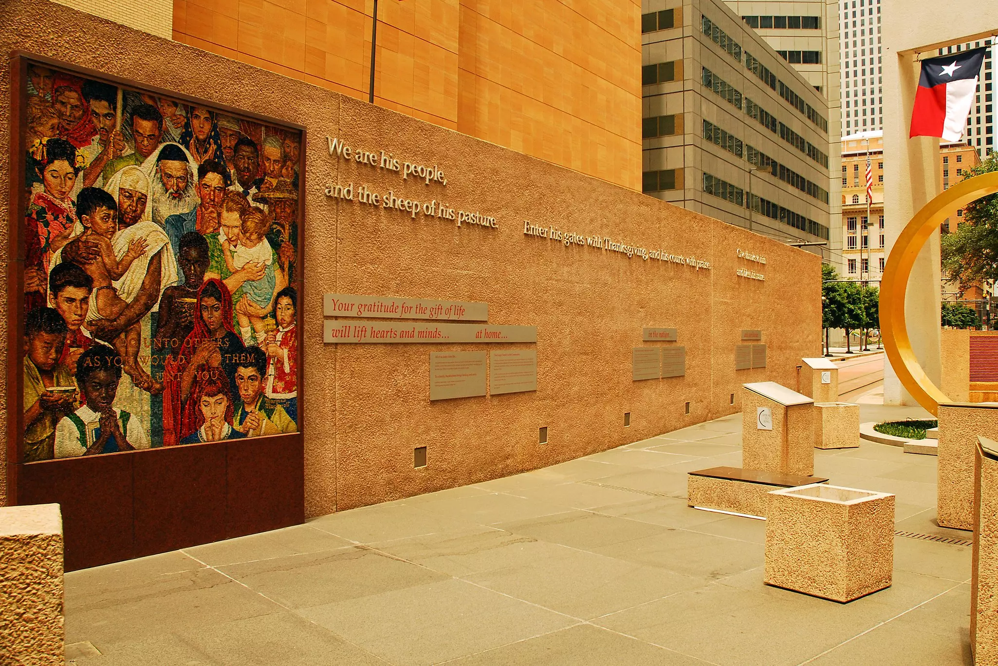 A city plaza with a mural of people celebrating different religions and words regarding gratitude and spirituality. The plaza also has stone sculptures and Texas state flag; city buildings are in the distance.