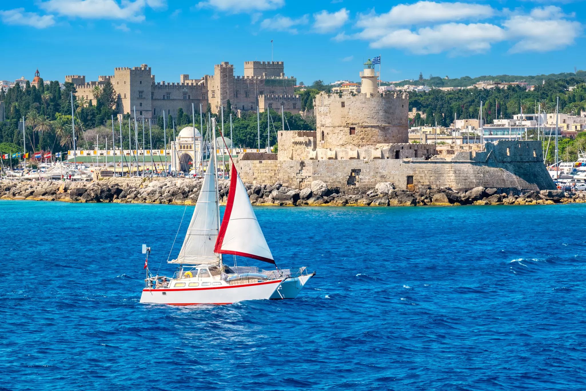 A red and white catamaran sailing past an old stone fortress on an island.
