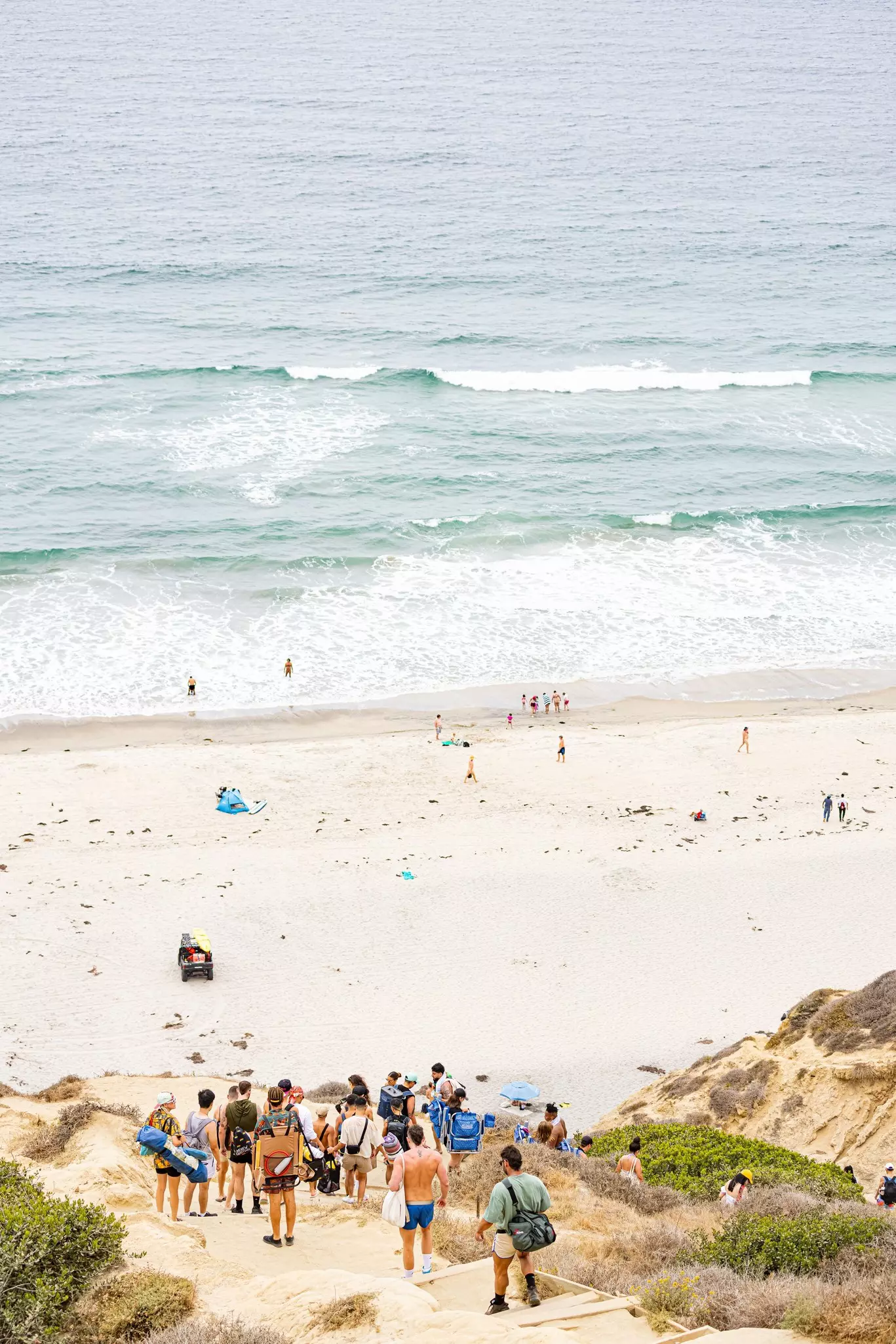 People carry bags and hike down a steep sandy cliff path towards the ocean.