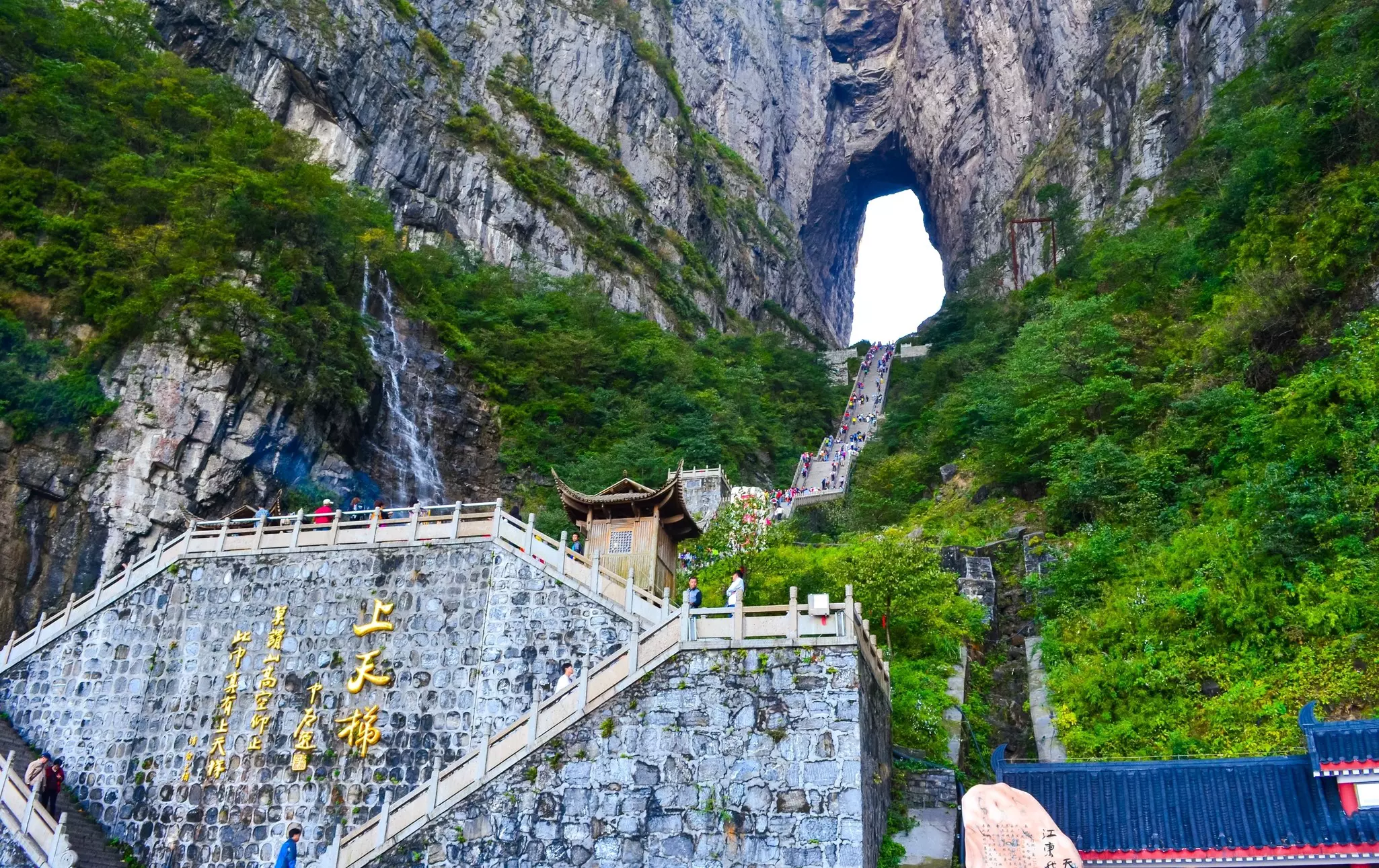 A long flight of stone steps lead from a landing inscribed with golden characters up to a small opening in a karst rock face. Verdant green vegetation covers the rocky slopes.