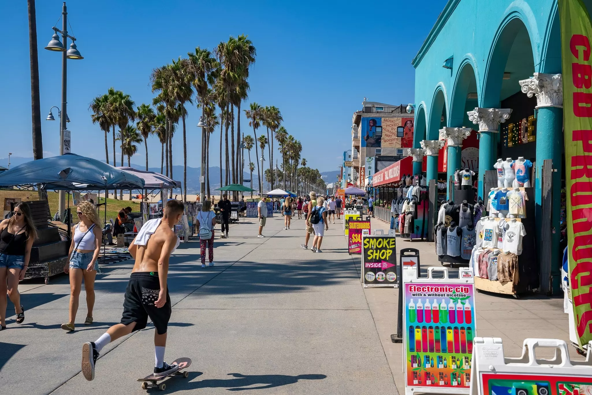 A skateboarder passes shops along a beachfront commercial strip. Other people walk along the path, and palm trees are visible beneath a blue sky.