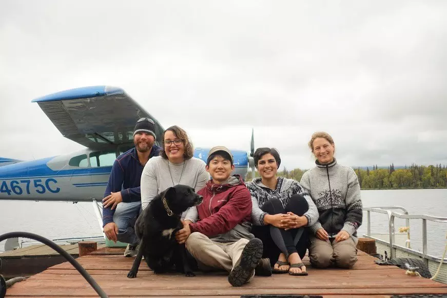 Five people and a dog sit on a dock with a seaplane in the background on an overcast day.