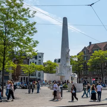 People crossing over Amsterdam's Dam Square, with the famous Nationaal Monument, a white obelisk, in the center on a sunny day.