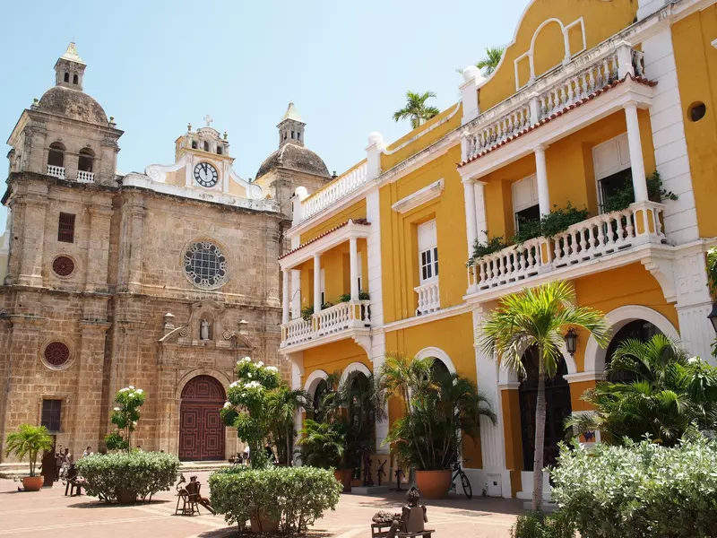 A tan stone church and a yellow-and-white building are positioned at one end of a plaza with tropical plants in the center.