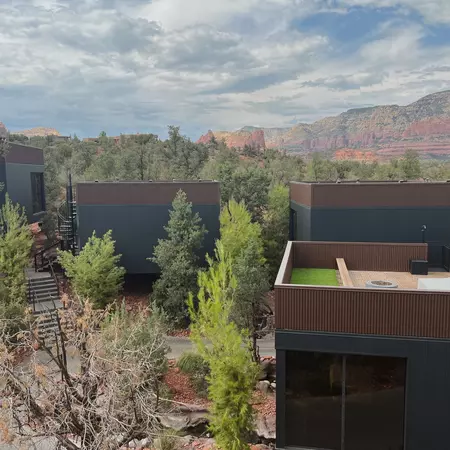 Structures surrounded by trees with reddish rock formations in the background under cloudy skies.