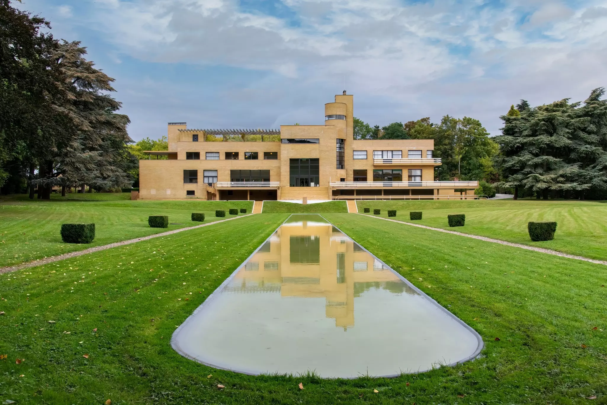 A large art deco-style building with many rectangular features and one rounded tower faces a large lawn with a central oval water feature.