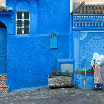 Woman in a white headscarf and striped robe at a blue carved fountain beside a blue door in Chefchaouen's medina.