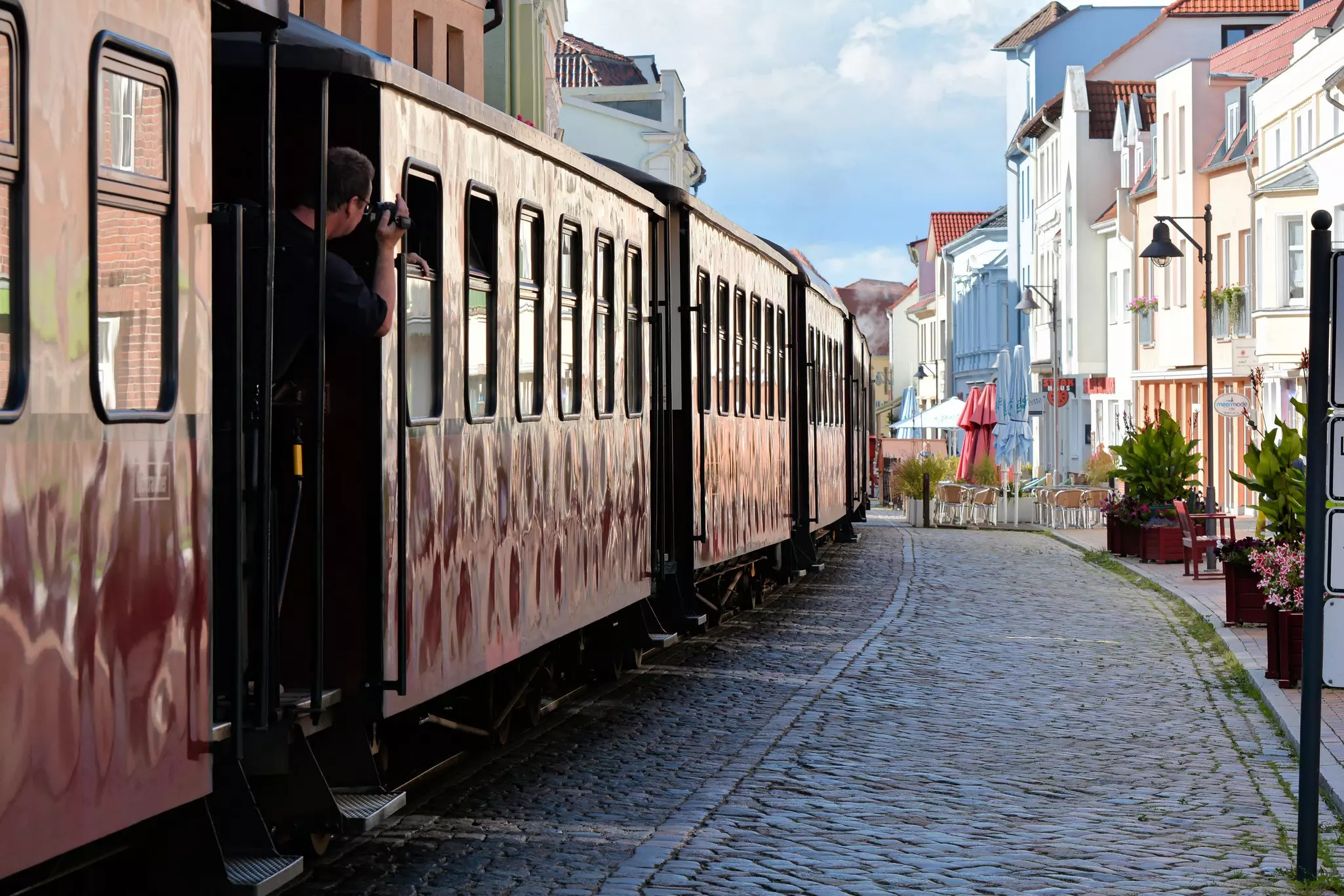 A man slightly leans out of a train to take a photo of an old cobbled stone town