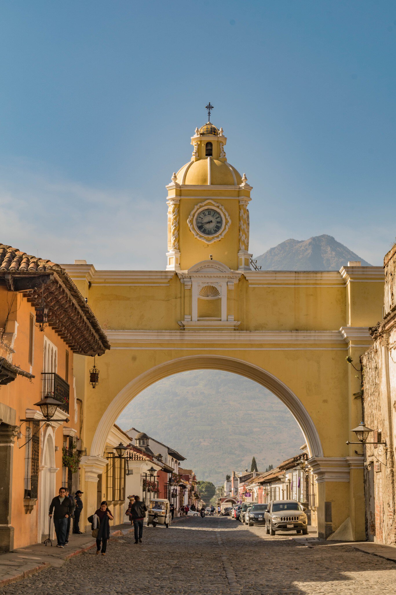 Yellow arch topped by a clock tower and bell cupola over a cobbled lane lined with colonial houses, mountain beyond