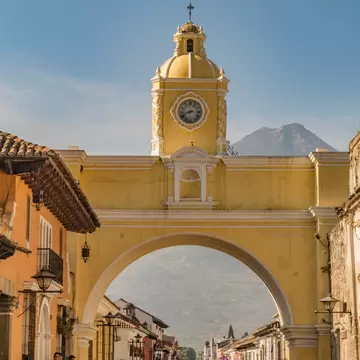 Yellow arch topped by a clock tower and bell cupola over a cobbled lane lined with colonial houses, mountain beyond