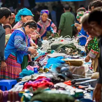 People in colorful, traditional outfits trade dry goods around a table at an outdoor market in Vietnam.