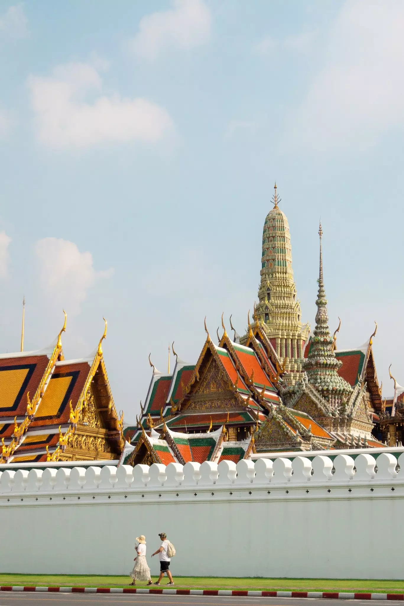 A couple walks in front of a white wall, behind which rises the elaborate roofs of a Buddhist temple complex.