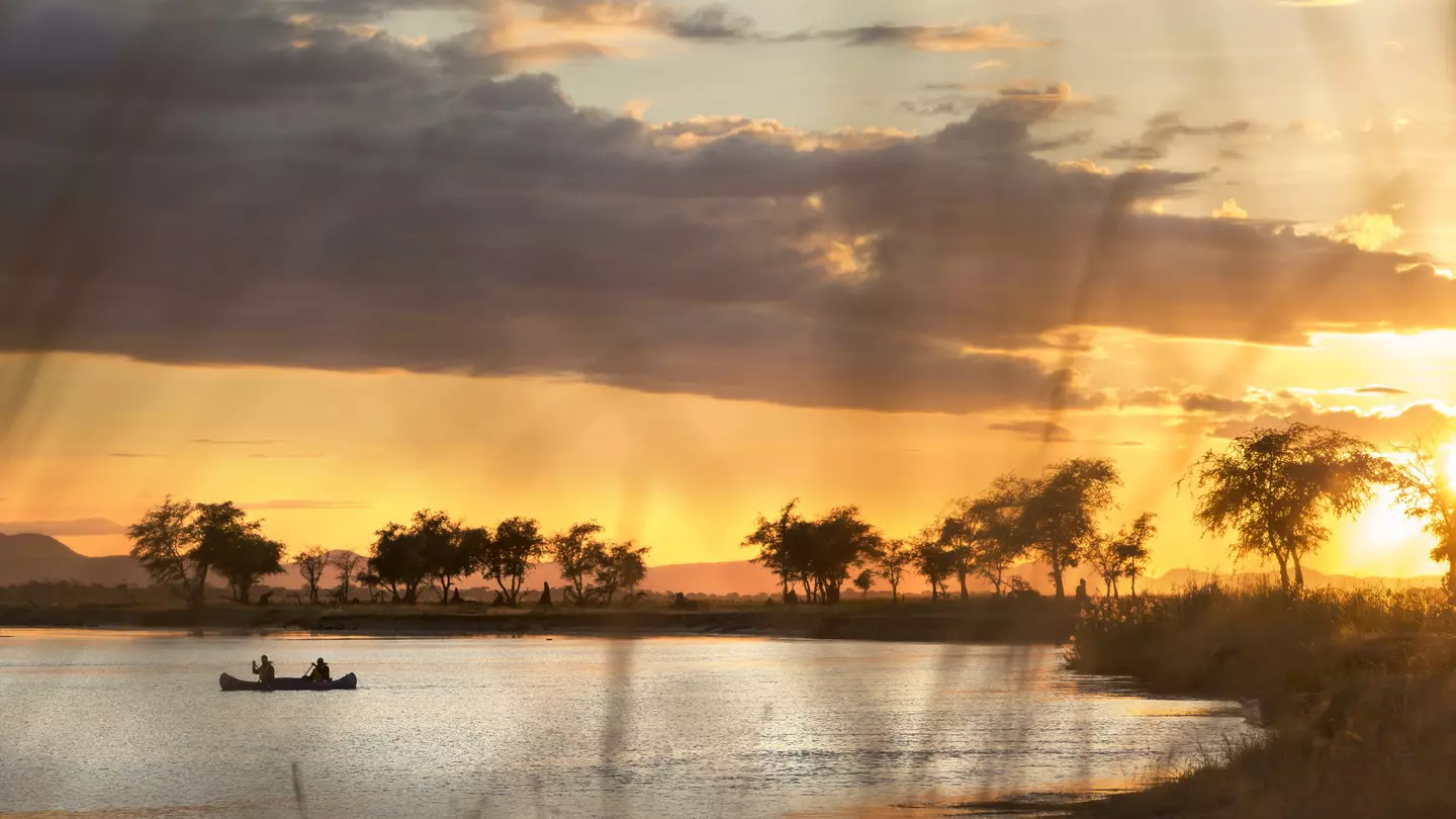 A canoe sits in a curve of the Zambezi River, with acacia trees silhouetted along its shore; the sky is golden, with dark clouds cutting across it.