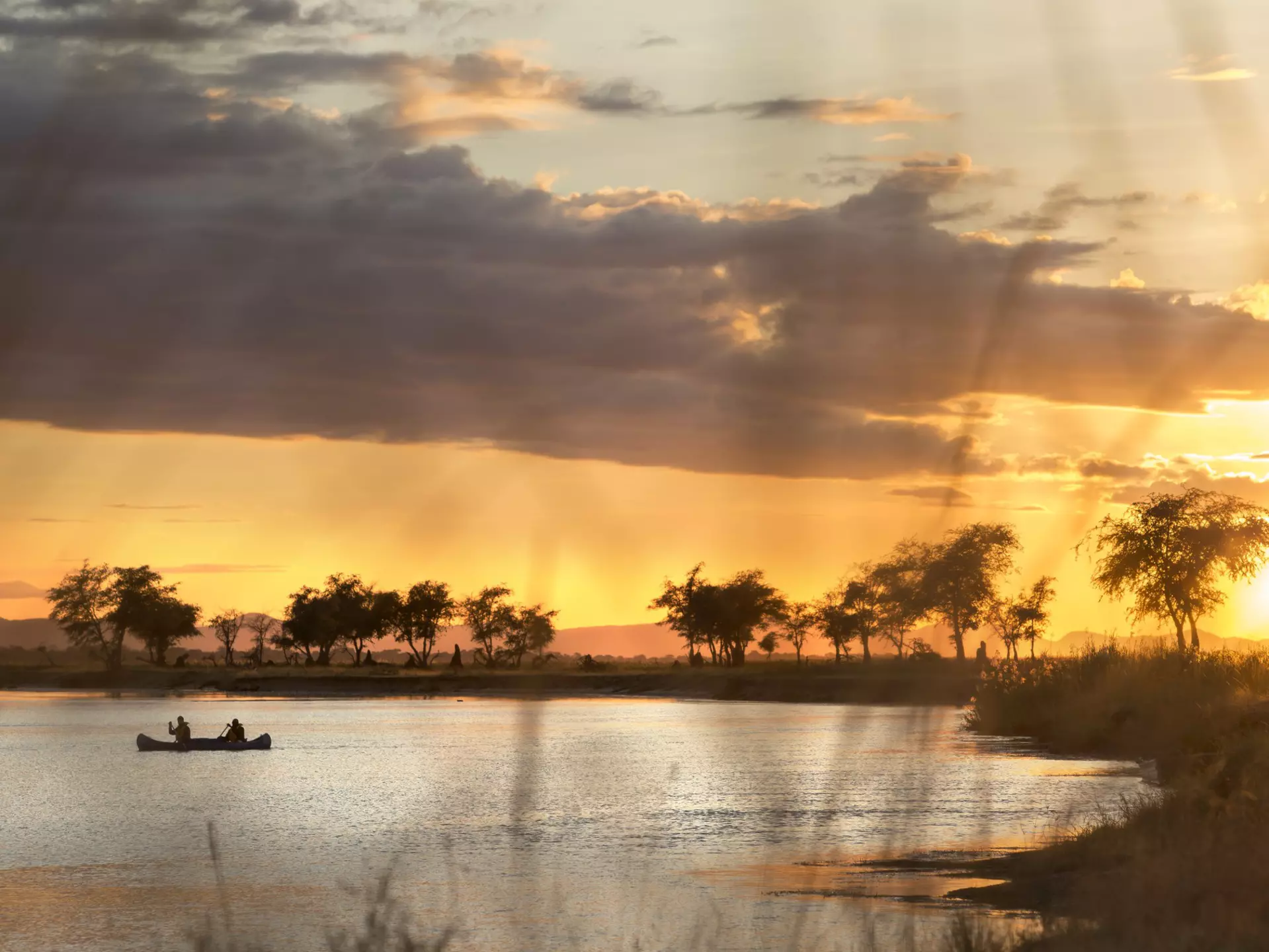 A canoe sits in a curve of the Zambezi River, with acacia trees silhouetted along its shore; the sky is golden, with dark clouds cutting across it.