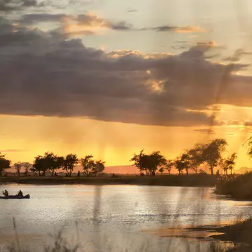 A canoe sits in a curve of the Zambezi River, with acacia trees silhouetted along its shore; the sky is golden, with dark clouds cutting across it.