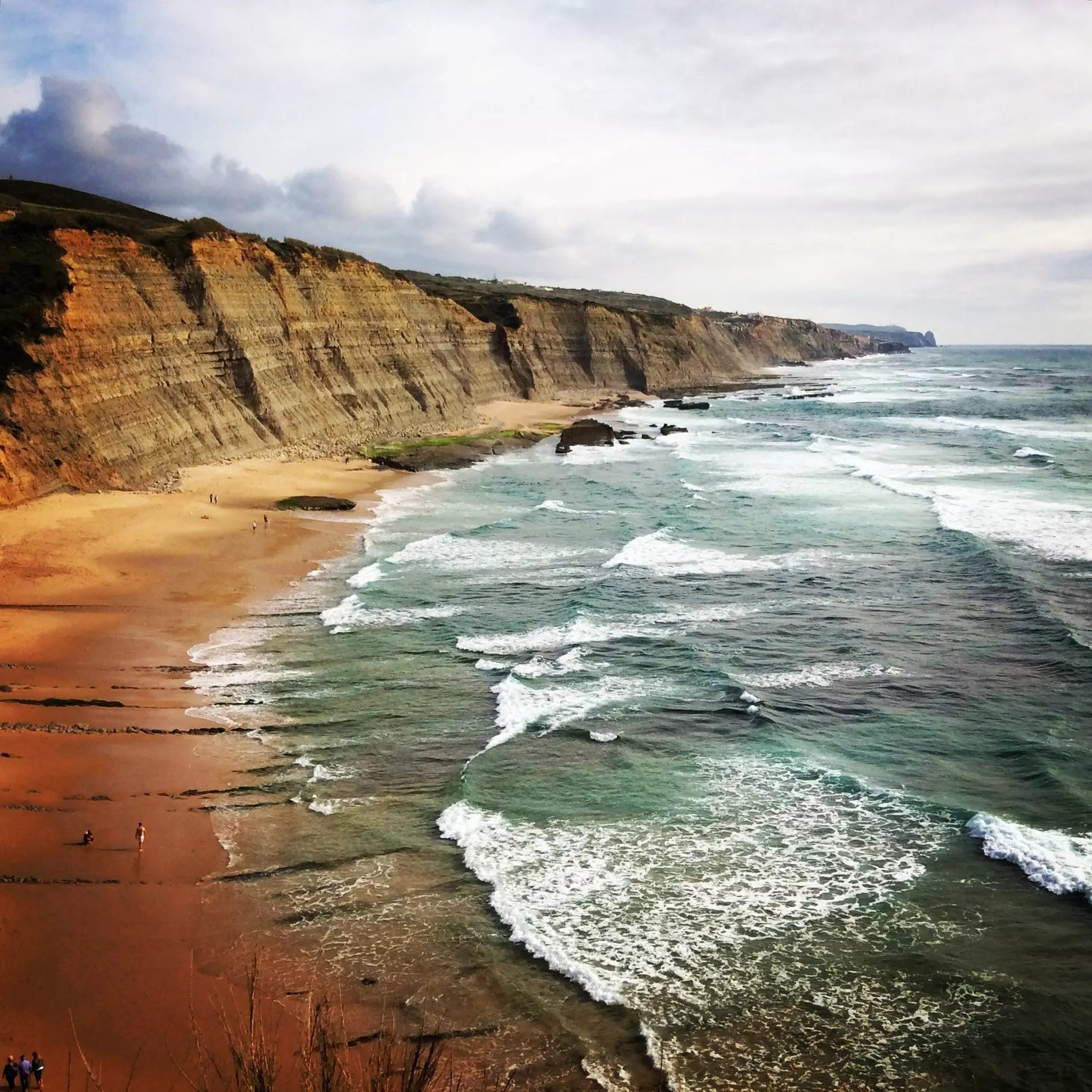 Waves crash on a sandy beach backed by sloping cliffs