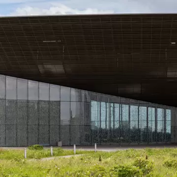 Long, low glass building with a huge dark cantilevered roof rising to one side, fronted by wild grass and small bollards