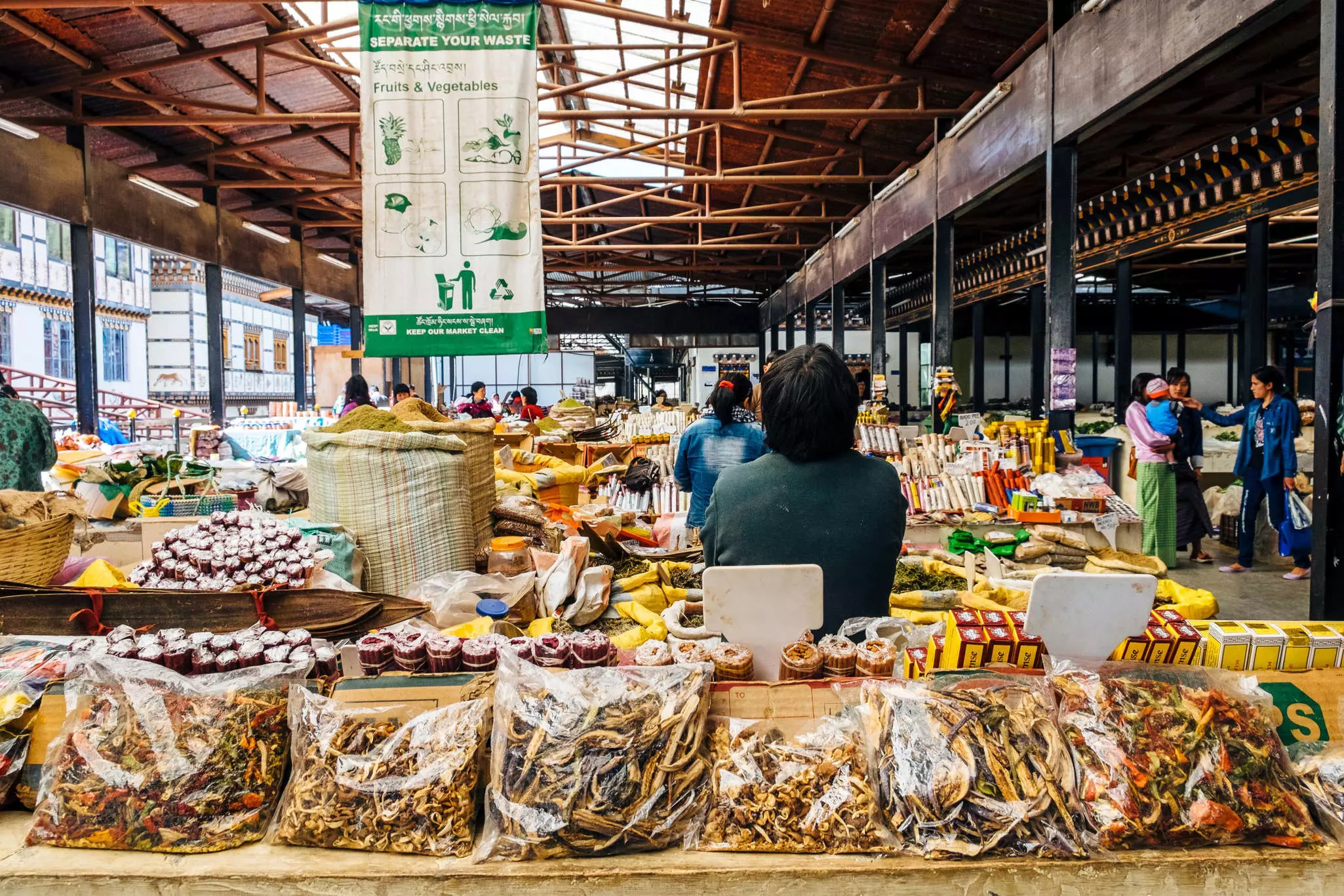 A woman is seen from behind at a market stall with dried vegetables and spices for sale.