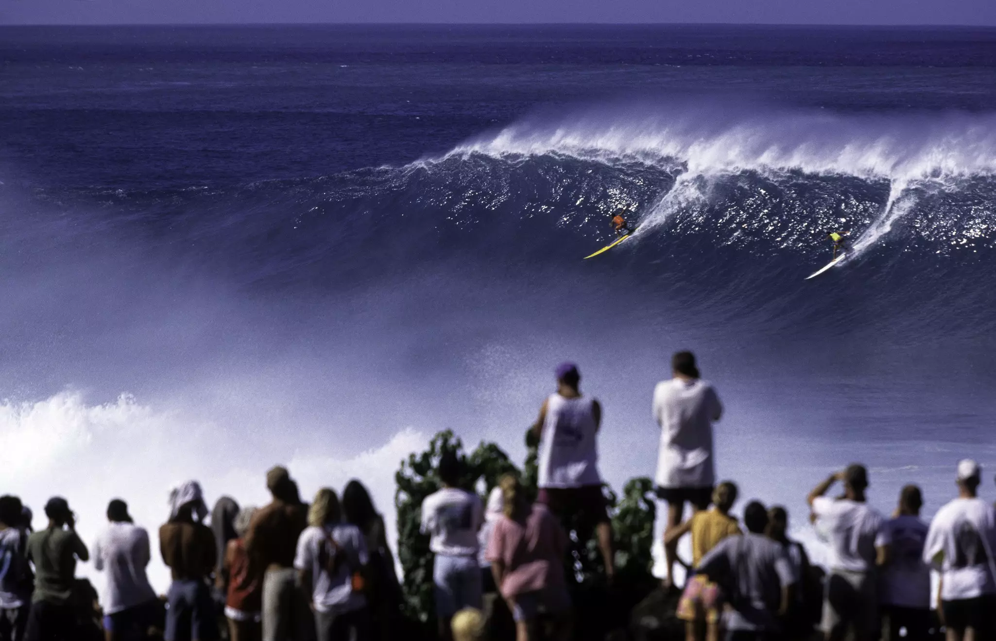 A crowd of onlookers in the foreground watch two surfers at the very top of a giant wave.