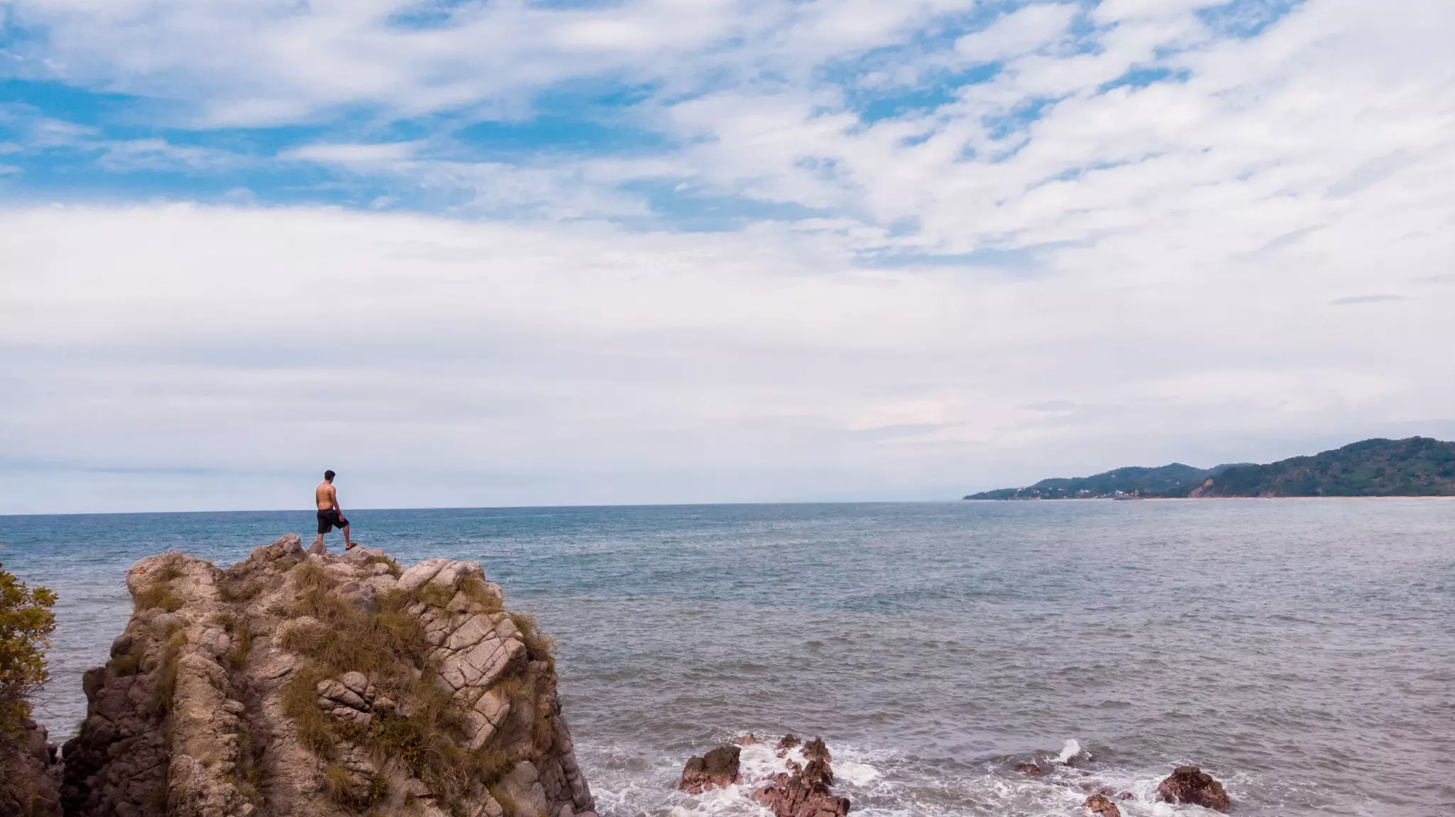 A man stands on a large rocky outcropping admiring the ocean along a jungly coastline