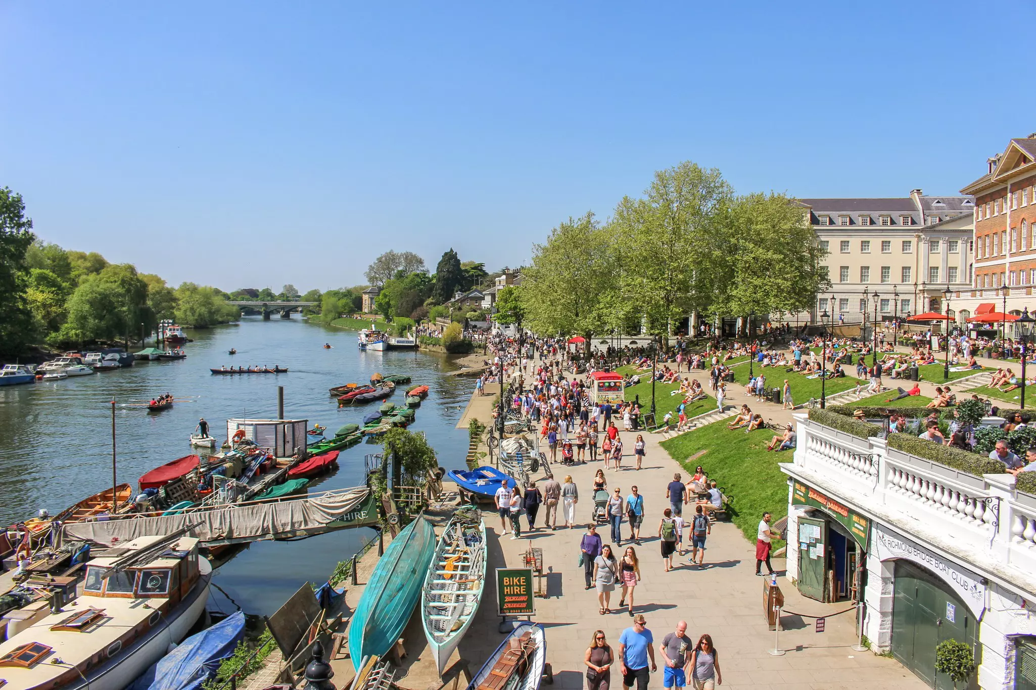 People walk beside a river on a sunny day. Rowers are out in boats and many people are sat relaxing on nearby lawns.
