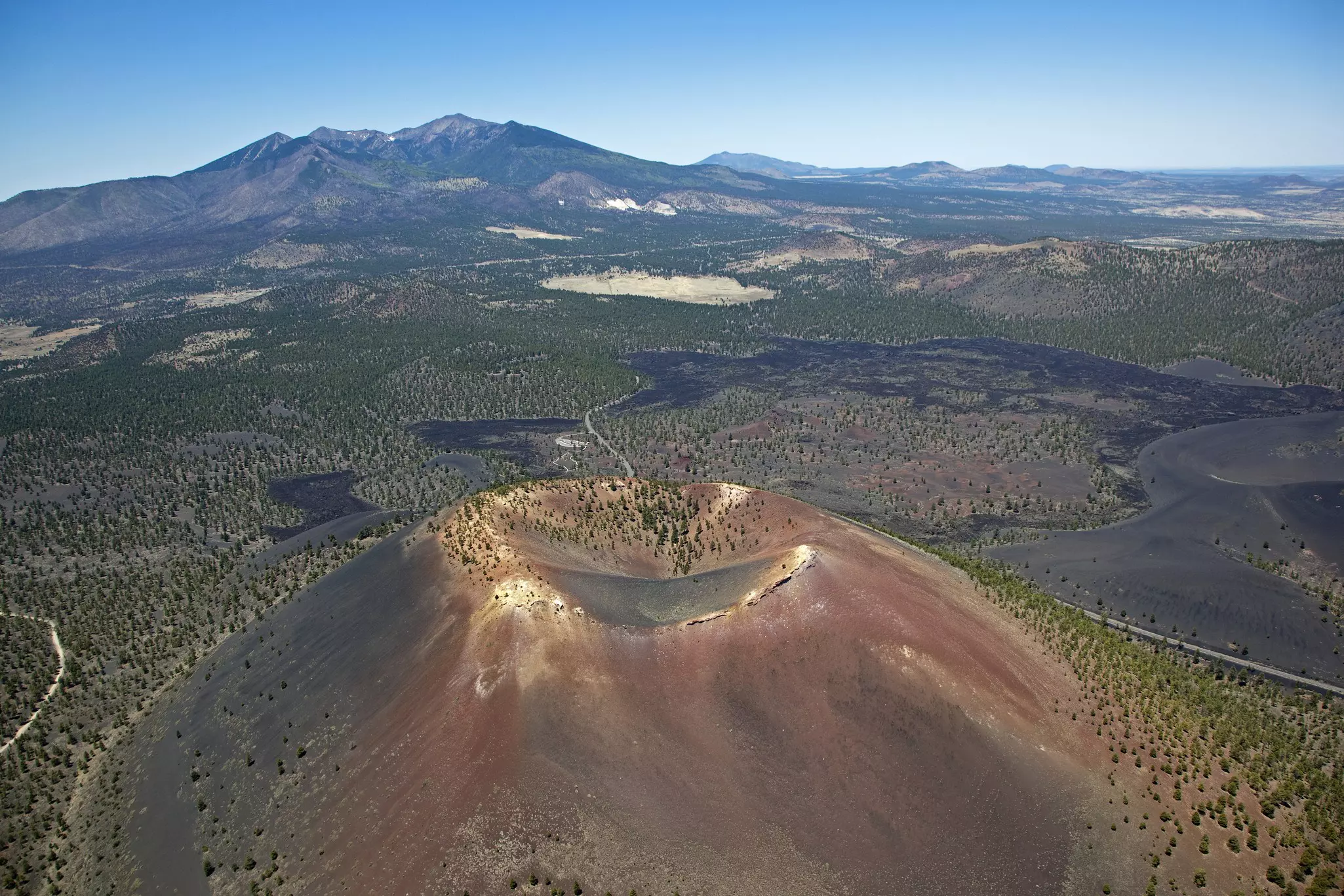 Aerial view of a volcanic crater with sparse forests and mountains in the distance on a sunny day.
