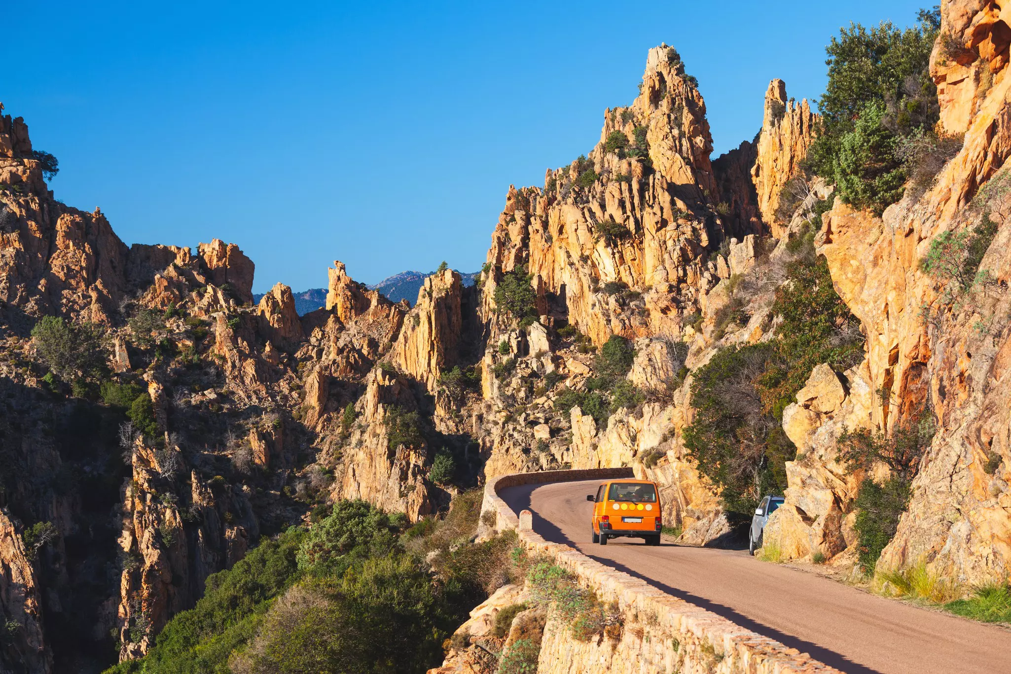 An orange van drives on a narrow road through rocky mountain outcrops.