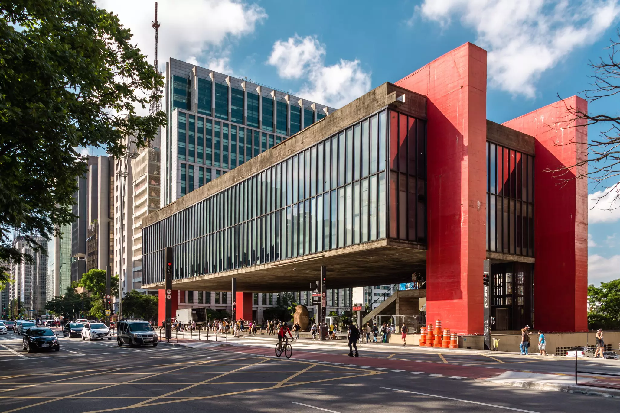 A large contemporary building with floor-to-ceiling glass windows supported by large red pillars appears to float above the street.