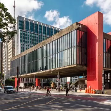 A large contemporary building with floor-to-ceiling glass windows supported by large red pillars appears to float above the street.