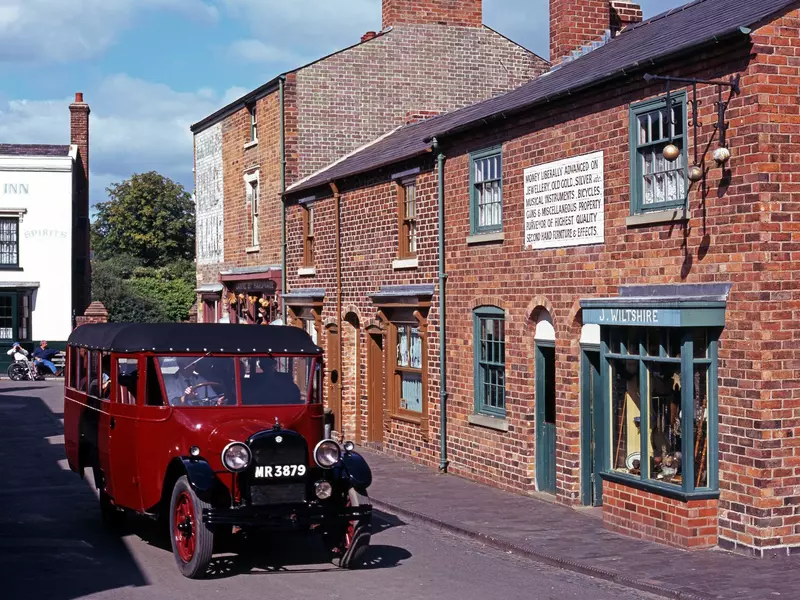 A red vintage car being driven along a recreated historic street of redbrick terraced homes and shops.