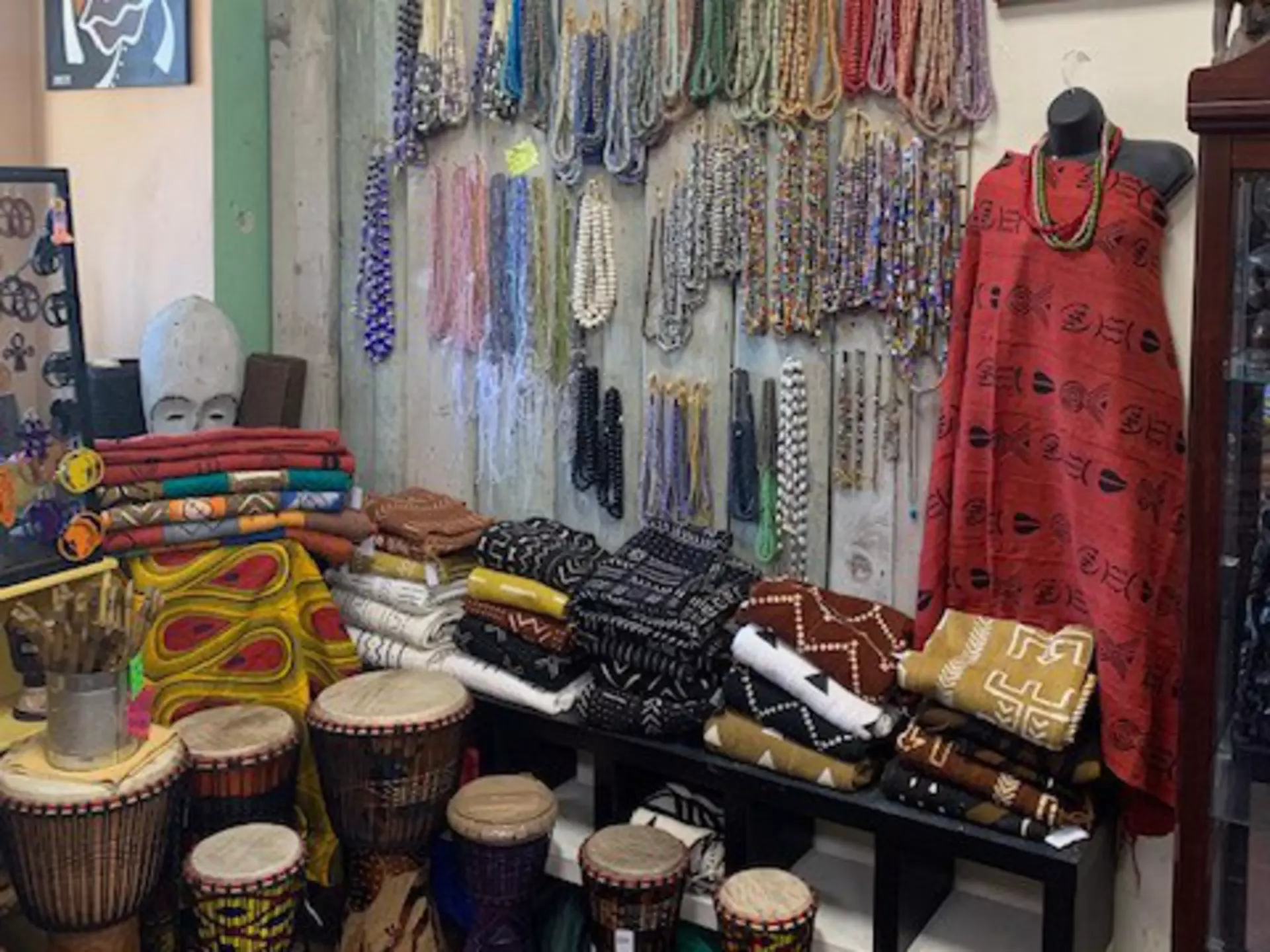 Interior shot of the Sika Jewelry store. There is a large display of necklaces hanging from the wall. Folded fabric and clothing placed on a wooden shelf. A headless mannequin is wearing draped in fabric and there are small drums placed on the floor
