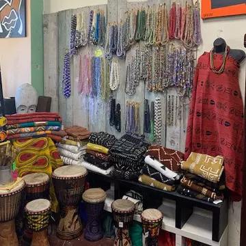 Interior shot of the Sika Jewelry store. There is a large display of necklaces hanging from the wall. Folded fabric and clothing placed on a wooden shelf. A headless mannequin is wearing draped in fabric and there are small drums placed on the floor