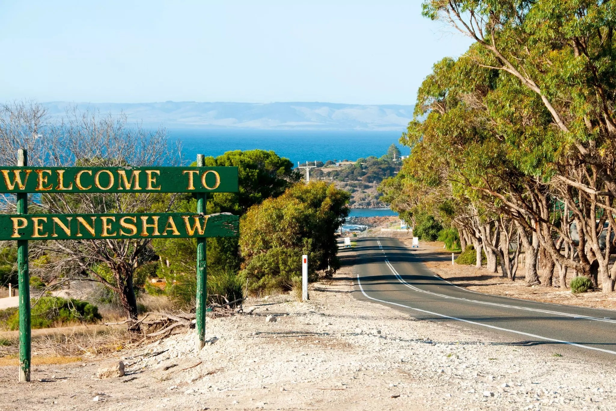 A road leads down to a small harbor town. A large road sign says "Welcome to Penneshaw".