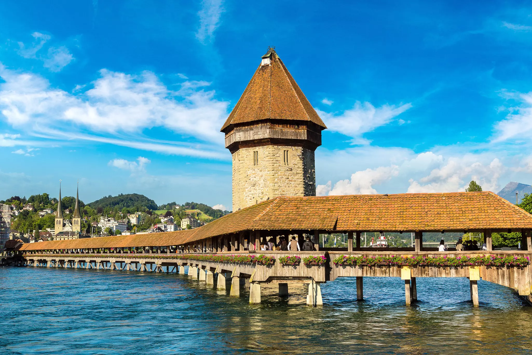 A covered wooden footbridge over a lake. The bridge is lined with flowerbeds.