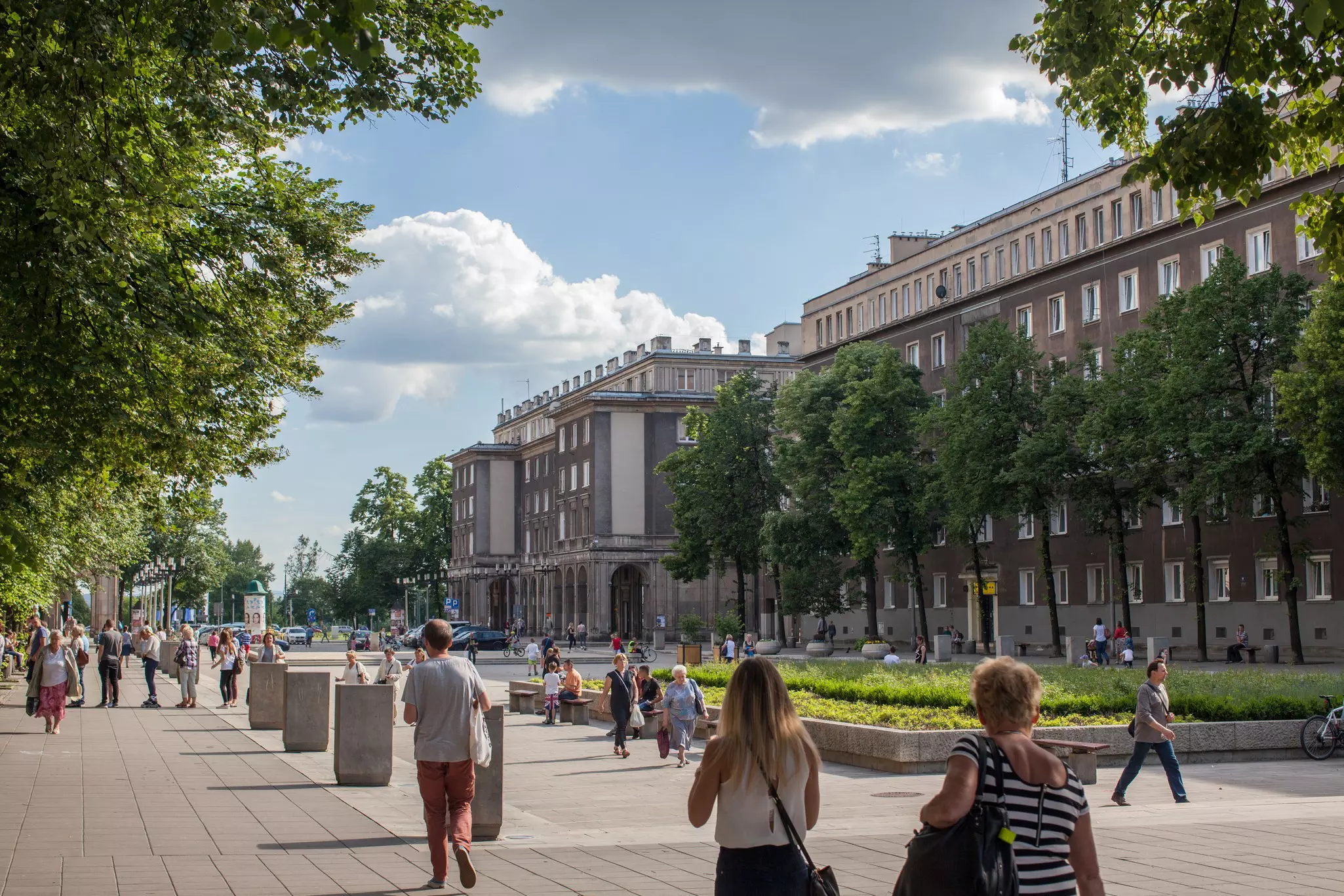 People walking down a broad pedestrian avenue with gray buildings to one side and a large raised bed of green plantings.