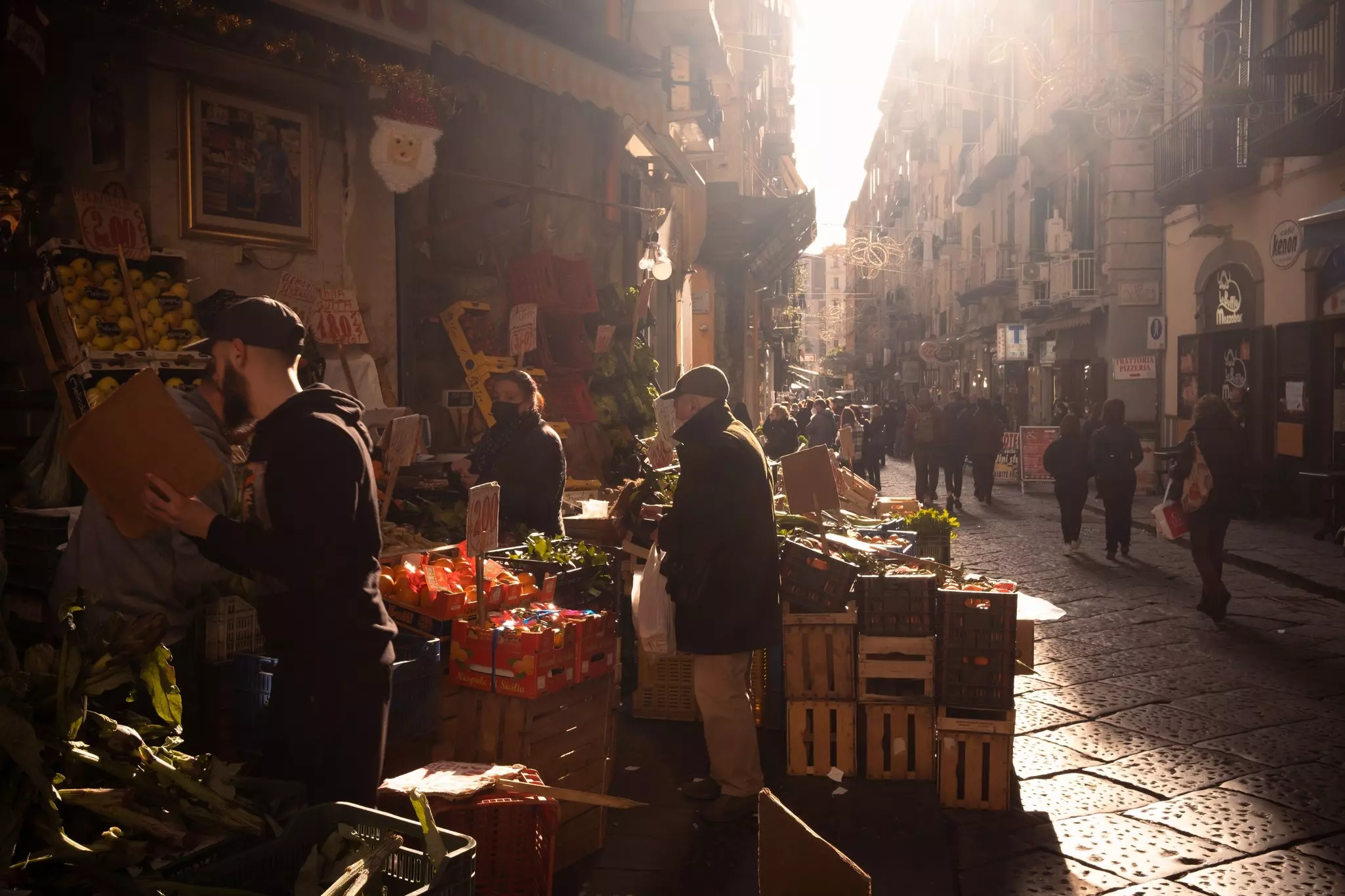 Vendors sell fresh vegetables in an open air market in a historic city center as people walk by the nearby street. The early morning light gives the scene a dramatic feel