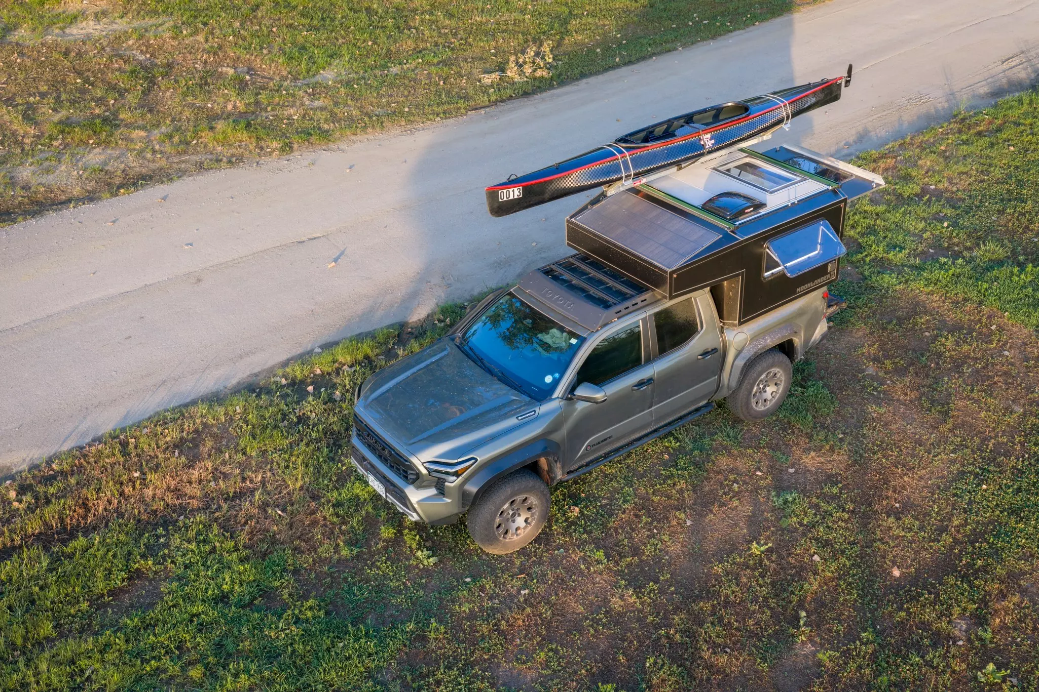 Aerial shot of a gray pickup on a roadside. A camper with a kayak on top is attached to the truck bed.