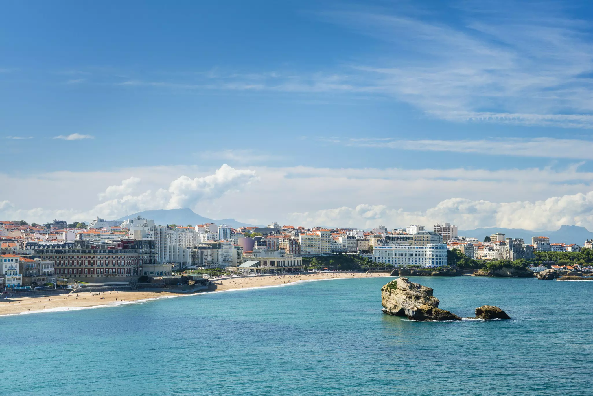 A large beach backed by a town with large buildings. A big rock sits in the middle of the shoreline