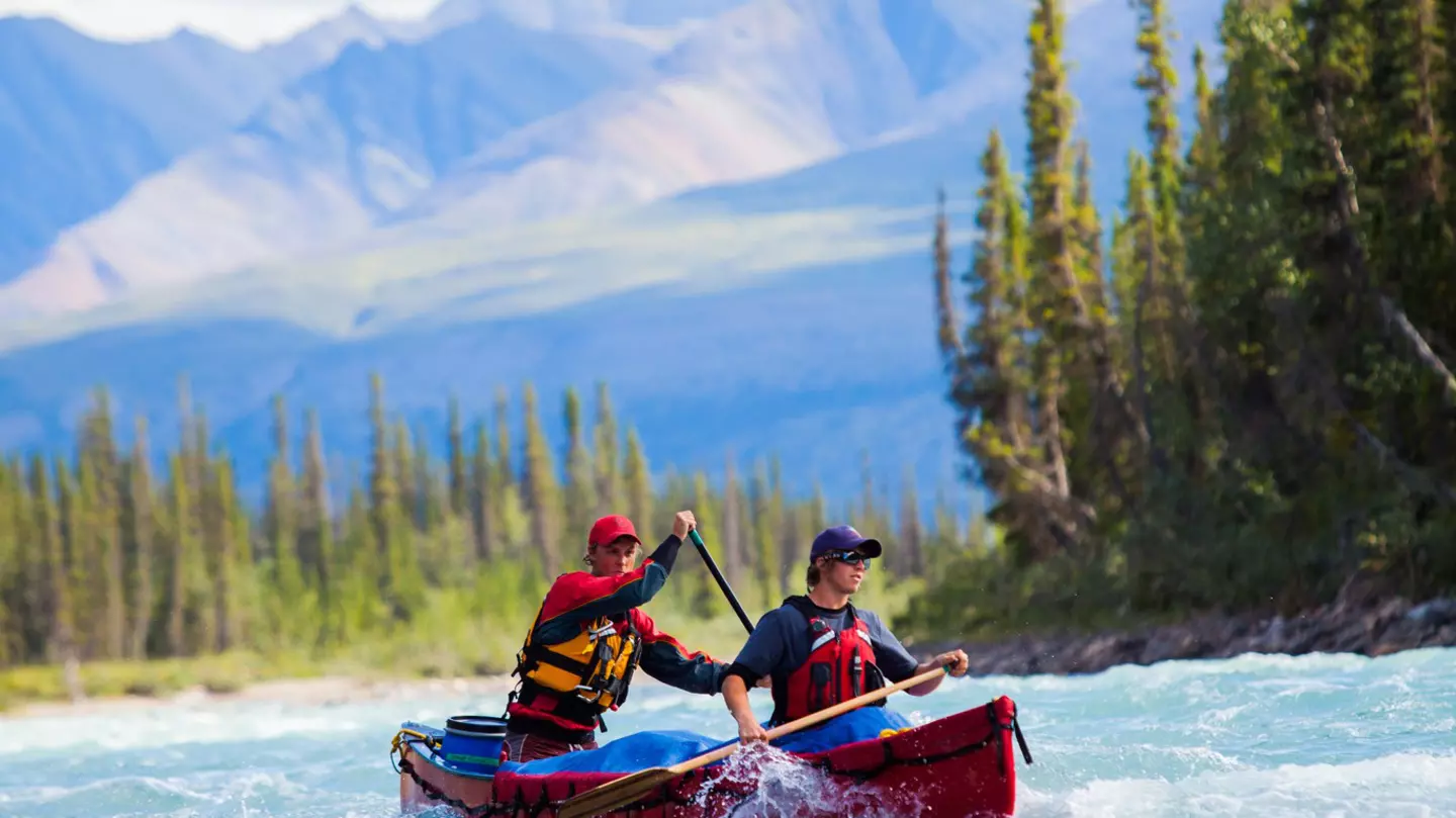Two men in a red boat paddling on a choppy river, with mountains in the blurred background and rocks in the foreground.
