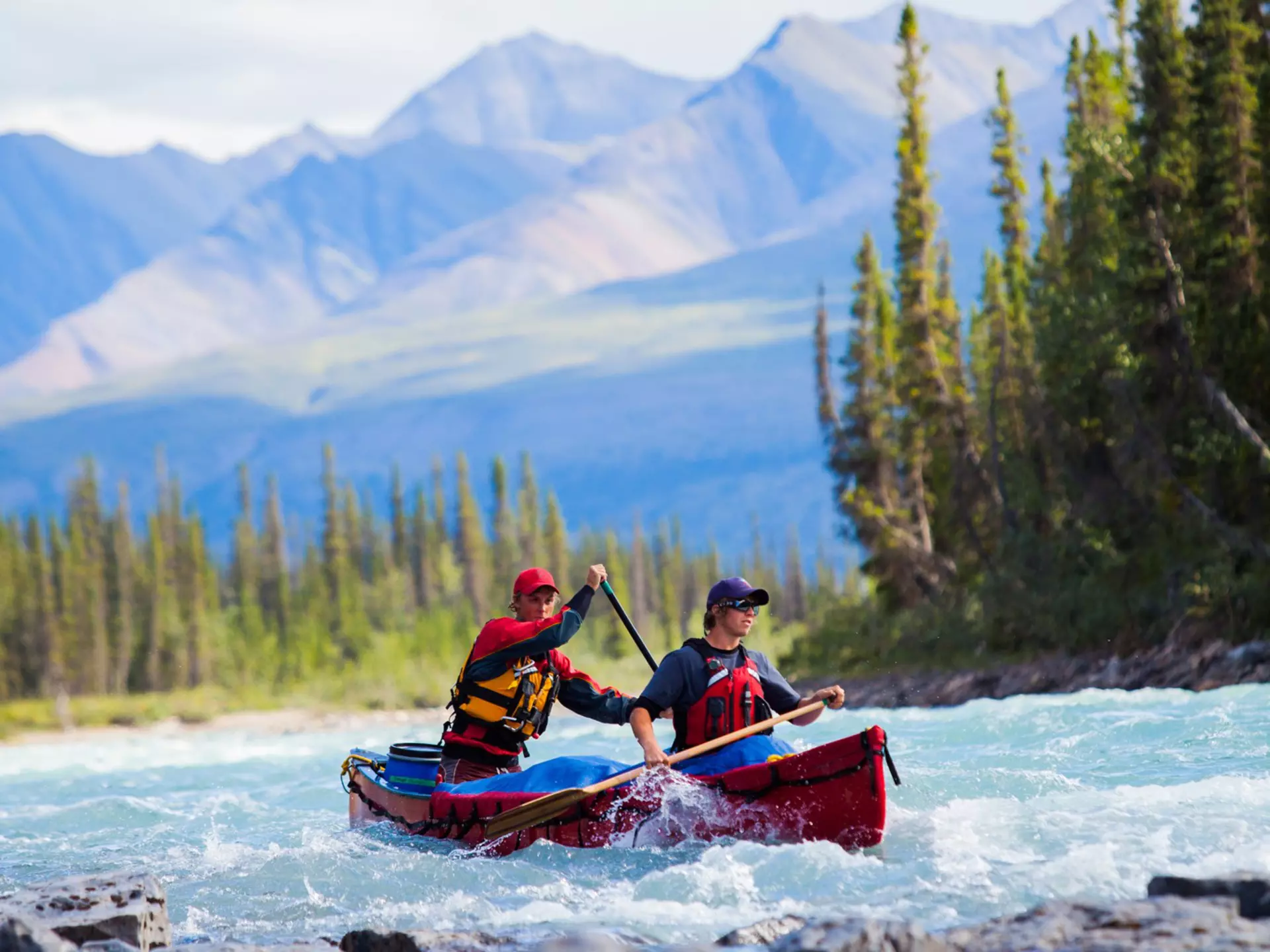 Two men in a red boat paddling on a choppy river, with mountains in the blurred background and rocks in the foreground.