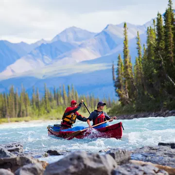 Two men in a red boat paddling on a choppy river, with mountains in the blurred background and rocks in the foreground.