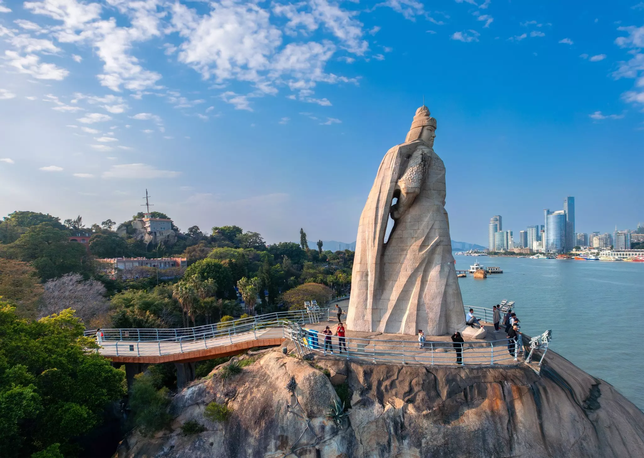 A walkway leads to a giant statue on a rock facing the water in Gulangyu China; people are standing by the railing surrounding the statue.
